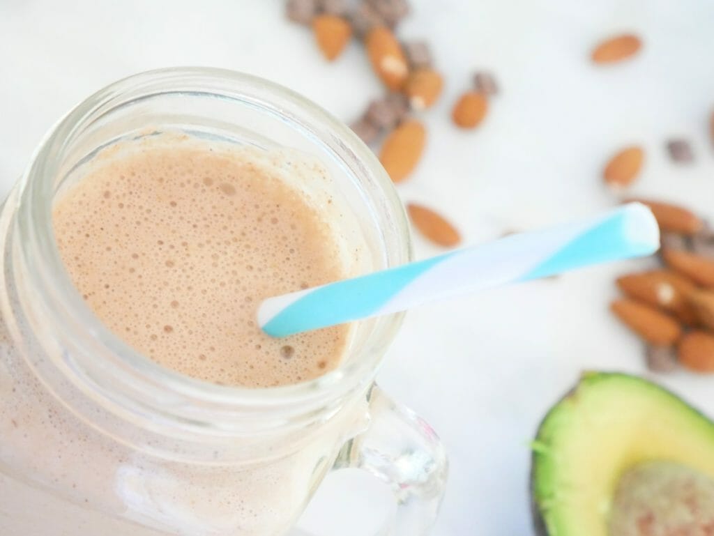 An almond butter protein shake with chocolate from above with avocado and almonds around and a blue straw