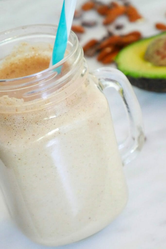 A chocolate and almond butter smoothie with coconut and protein with avocado and almonds behind