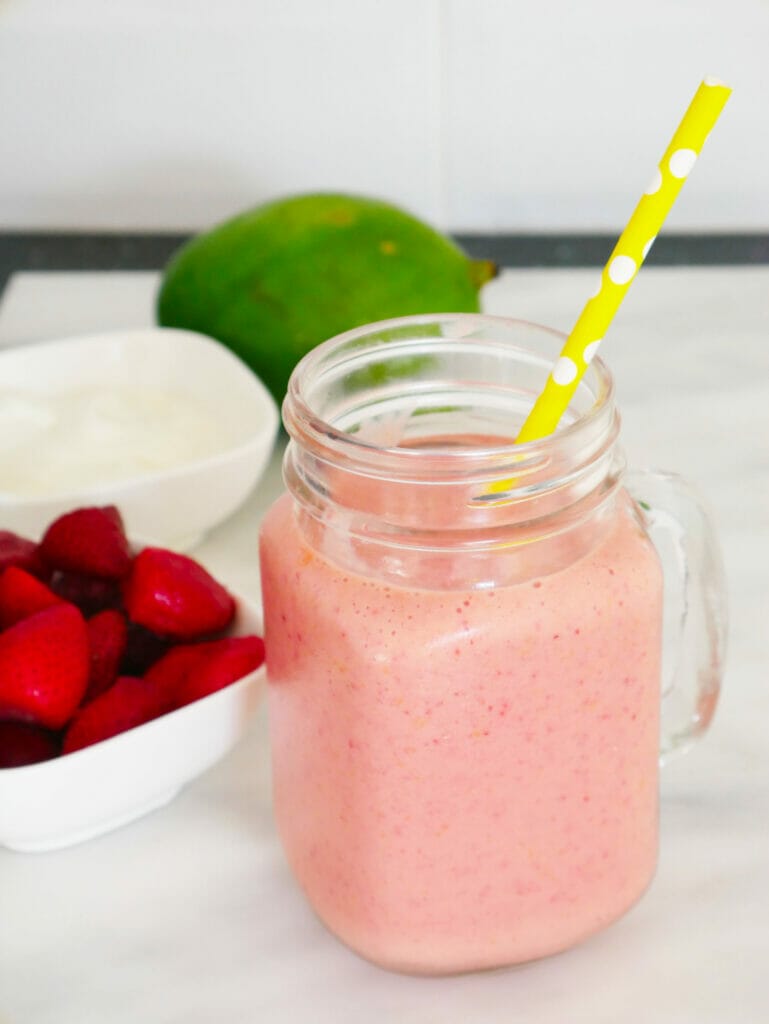 Strawberry mango smoothie with yellow straw and recipe ingredients behind
